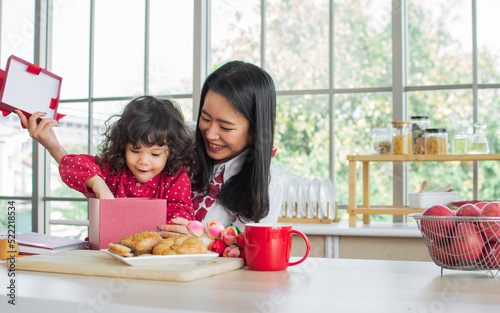 Beautiful Asian mother giving gift box to mixed race adorable little daughter, sitting in kitchen at home to celebrate birthday or Valentine day together, smiling and surprising with happiness.