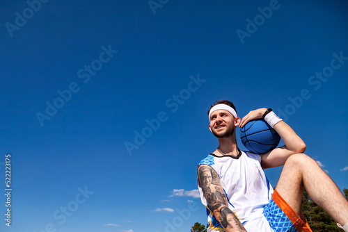 Sports and basketball concept. Young man with basketball ball in arm looking away smiling at blue sky outdoors.