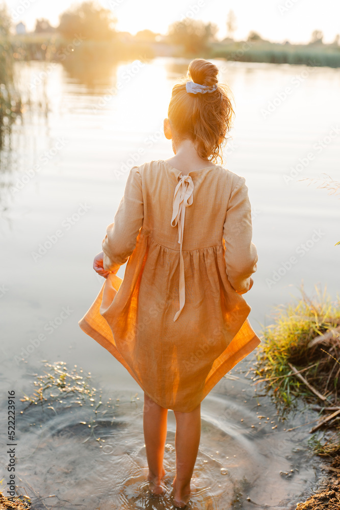 Φωτογραφία Curly caucasian girl wearing natural linen dress standing turned back in lake water, summertime