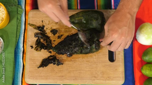 Overhead shot of a roasted poblano pepper is peeled with the edge of a spoon on a wooden cutting board on a colorful Mexican mantel piece. This vegetable is a delicious staple in Mexican cuisine. photo