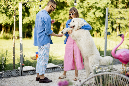 Young stylish couple hang out together and play with a dog at their beautiful backyard of country house. Man and woman spend summertime with pet outdoors
