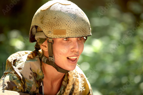 Caucasian Military lady woman in tactical gear posing for photo at summer season. Wearing green camo uniform and assault rifle, in military gear and headset, lady is looking at side