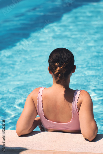 Young Caucasian woman in bikini, sitting relaxed on the shore of a crystal clear pool. Concept holiday, travelling, vertical shot.