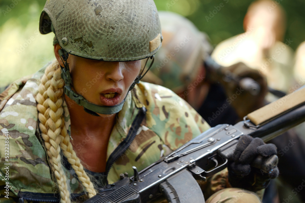 Caucasian Military lady woman in tactical gear posing for photo at summer season. Wearing green camo uniform and assault rifle, in military gear and headset, lady is looking at side