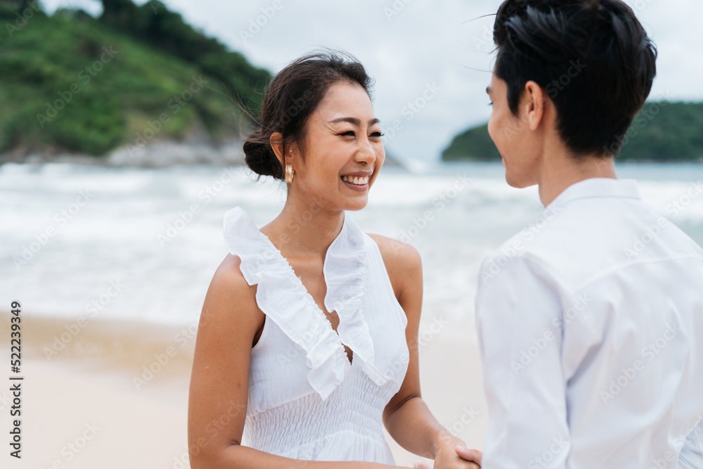 Obraz premium Happy toothy smiling Asian 30s couple in relationship in candid on the beach in summer. A romantic couple wearing a white shirt and dress.