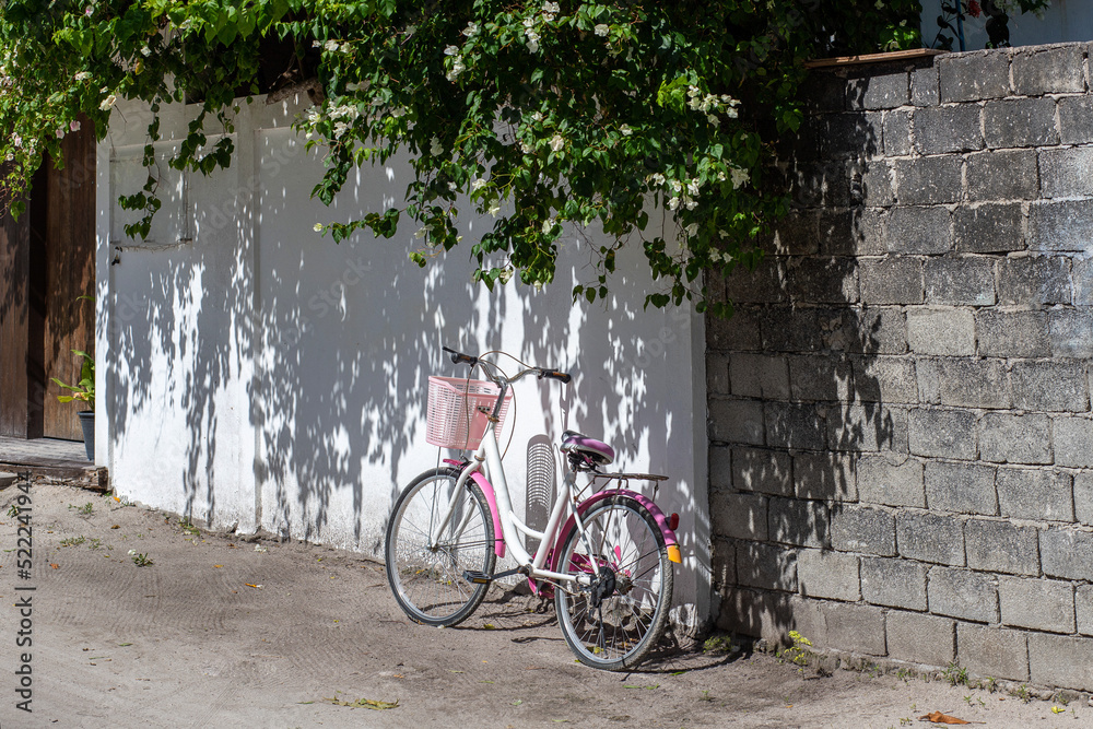 A white and pink retro bicycle with a basket stands near a white wall on the street