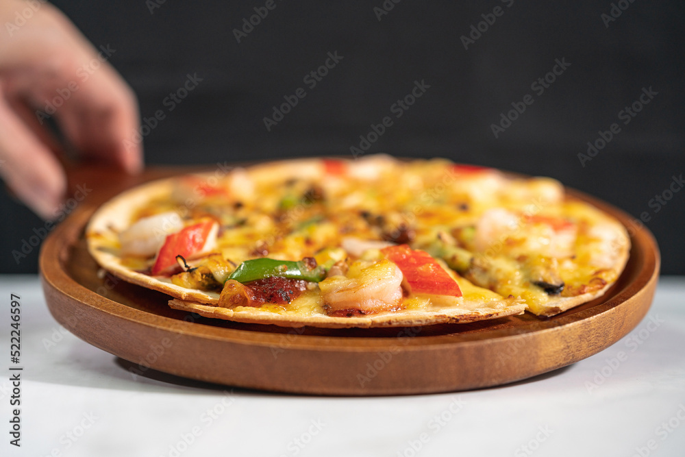 Hand served tasty hot baked seafood crispy pizza - mussels, shrimps and kani with chili pepper and melted mozzarella cheese on round pizza dough, on a wooden pizza pan over the marble table top.