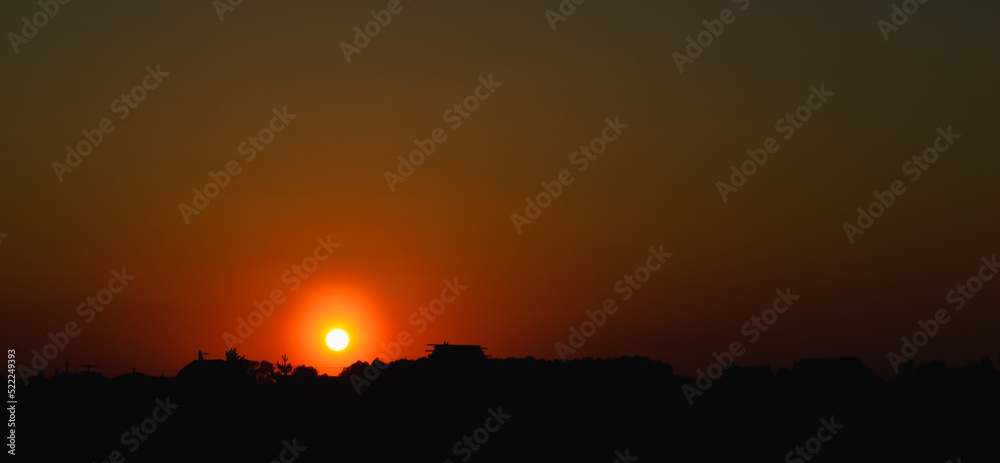 The setting sun at sunset in the orange evening sky above the silhouettes of buildings and trees The setting sun at sunset in the orange evening sky above the silhouettes of buildings and trees