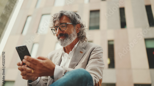 Mature businessman with beard in eyeglasses wearing gray jacket is using cell phone. Middle aged manager scrolling information on his smartphone while sitting outside the office