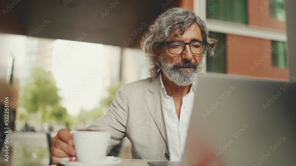 Mature businessman with beard in eyeglasses wearing gray jacket sits on cafe. Middle aged manager successful man user on laptop pc computer sit at cafe outdoors