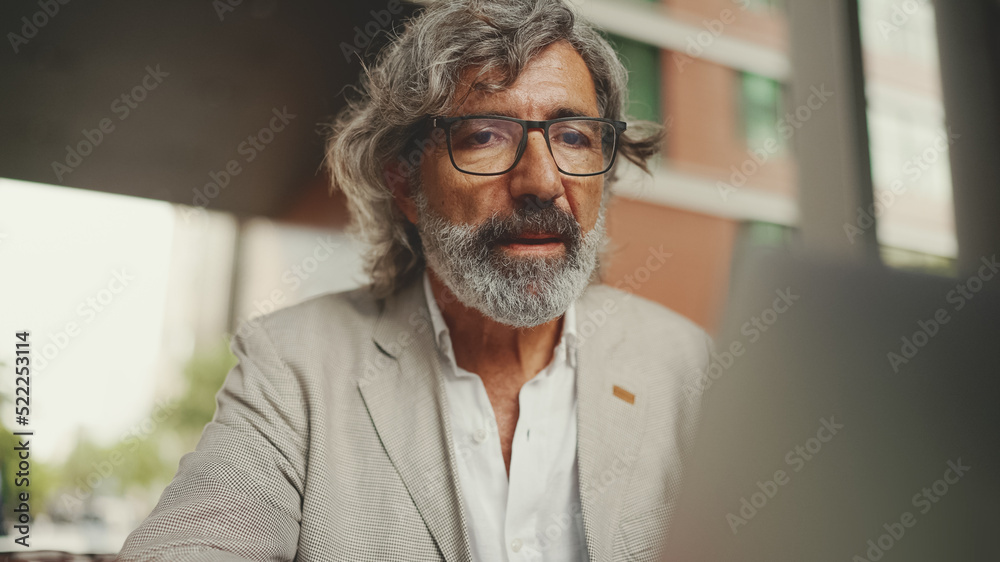 Clouse up, mature businessman with beard in eyeglasses wearing gray jacket sits on cafe. Middle aged manager successful man user on laptop pc computer sit at cafe outdoors Clouse up, mature businessman with beard in eyeglasses wearing gray jacket sits on cafe. Middle aged manager successful man user on laptop pc computer sit at cafe outdoors