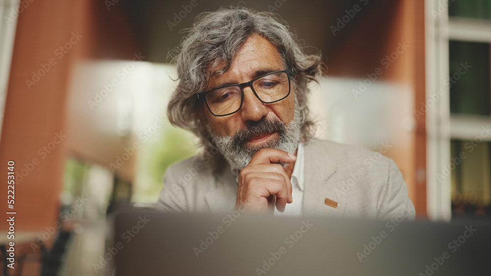 Fototapeta premium Clouse up, mature businessman with beard in eyeglasses wearing gray jacket sits on cafe. Middle aged manager successful man user on laptop pc computer sit at cafe outdoors