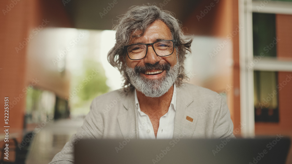 Mature businessman with beard in eyeglasses wearing gray jacket sits in cafe. Middle aged manager having video conference, video call on laptop pc computer sit at cafe outdoor Mature businessman with beard in eyeglasses wearing gray jacket sits in cafe. Middle aged manager having video conference, video call on laptop pc computer sit at cafe outdoor