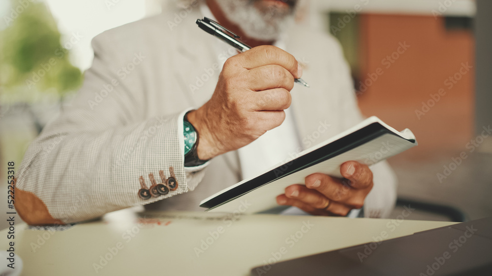 Fototapeta premium Mature businessman with beard in eyeglasses, user on laptop computer and taking notes, sitting in outdoor cafe. Successful man sitting at table in cafe in nature Mobile office freelancer