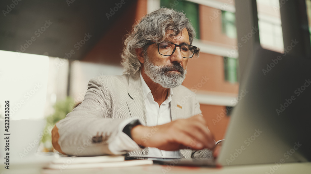 Mature businessman with beard in eyeglasses, user on laptop computer and taking notes, sitting in outdoor cafe. Successful man sitting at table in cafe in nature Mobile office freelancer Mature businessman with beard in eyeglasses, user on laptop computer and taking notes, sitting in outdoor cafe. Successful man sitting at table in cafe in nature Mobile office freelancer
