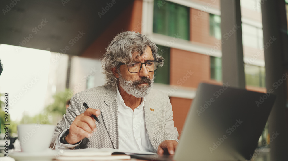 Mature businessman with beard in eyeglasses, user on laptop computer and taking notes, sitting in outdoor cafe. Successful man sitting at table in cafe in nature Mobile office freelancer