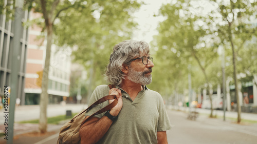 Middle-aged man with gray hair and beard walks and looks around. Mature gentleman in eyeglasses with bag on his shoulder walks through the square on the cityscape background