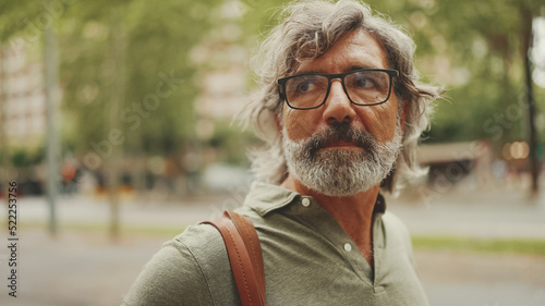 Middle-aged man with gray hair and beard walks and looks around. Mature gentleman in eyeglasses with bag on his shoulder walks through the square on the cityscape background