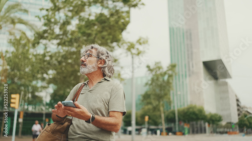 Friendly middle aged man with gray hair and beard looking at map trying to find his way using his mobile phone. Mature gentleman in eyeglasses using map app in cellphone outdoors © Andrii Nekrasov