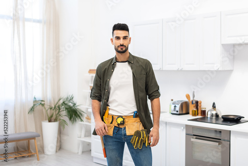 Arabian man in tool belt looking at camera in kitchen