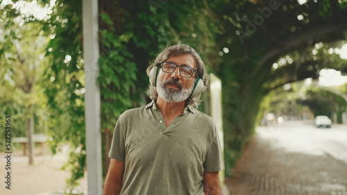 Friendly middle-aged man with gray hair and beard wearing casual clothes listening to music on headphones. Mature gentleman in eyeglasses enjoys music outdoors