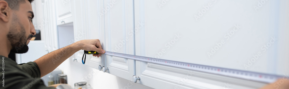 Side view of arabian man measuring cupboards in kitchen at home, banner