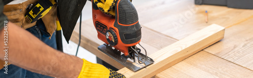 Cropped view of man in gloves using jigsaw machine on wooden plank at home, banner photo