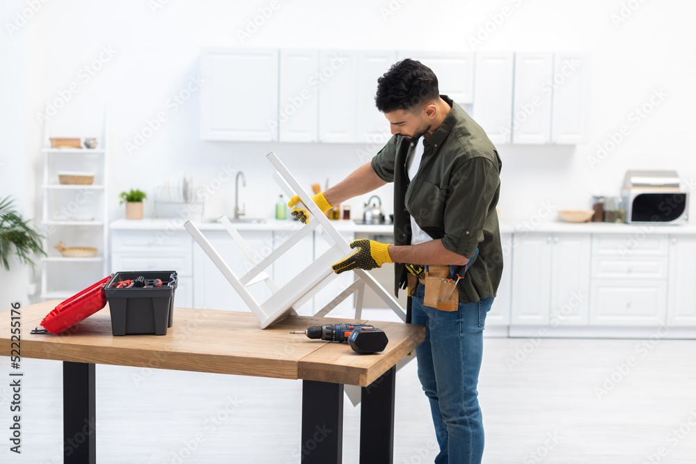 Arabian man with tool belt holding chair on table at home Arabian man with tool belt holding chair on table at home