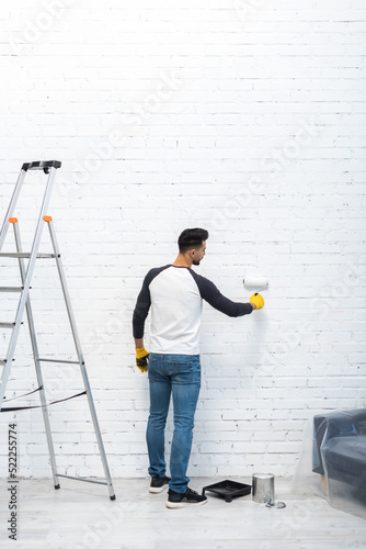 Side view of muslim man in gloves coloring brick wall with roller at home