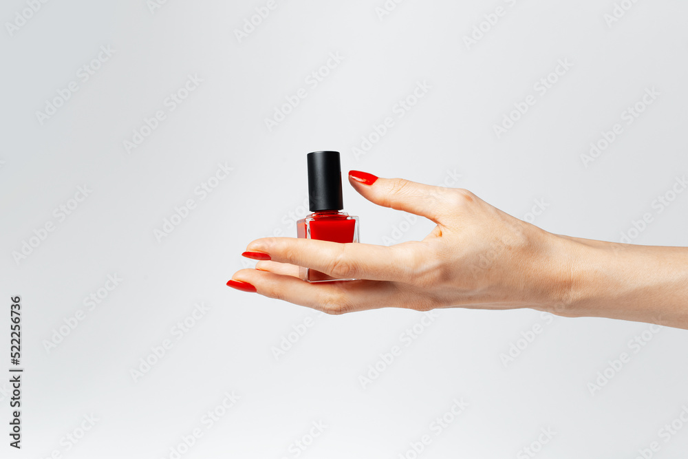 Fototapeta premium Close-up of female hand with red manicure holding a glitter nail polish of red color on white background.