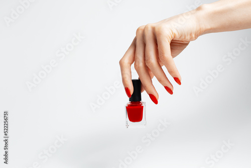 Close-up of female hand with red manicure holding a glitter nail polish of red color on white background.