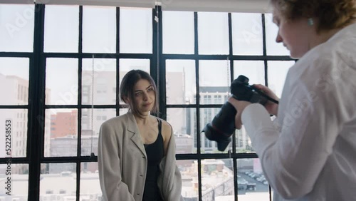 Motion footage backstage of professional photo session on DSLR camera in studio with natural daytime light on background. Young model arranging jacket, sitting next to window, posing, touching hair