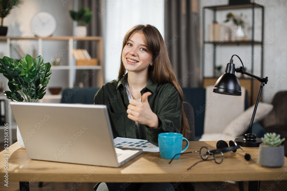 Beautiful caucasian woman with long dark hair sitting at desk and typing on laptop. Focused female in dark shirt and jeans using portable computer for work at bright office showing thumb up Beautiful caucasian woman with long dark hair sitting at desk and typing on laptop. Focused female in dark shirt and jeans using portable computer for work at bright office showing thumb up