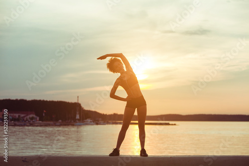 Attractive young African american girl silhouette exercising stretching on a beach after running at sunset