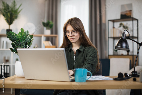 Beautiful caucasian woman in glasses with long dark hair working on wireless laptop while sitting at desk. Female freelancer in casual shirt using computer for remote work.