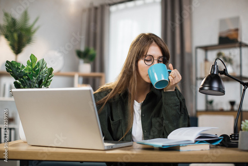 Young caucasian woman sitting at desk and surfing internet on portable computer while drinking coffee and reading the book. Concept of freelance and technology.