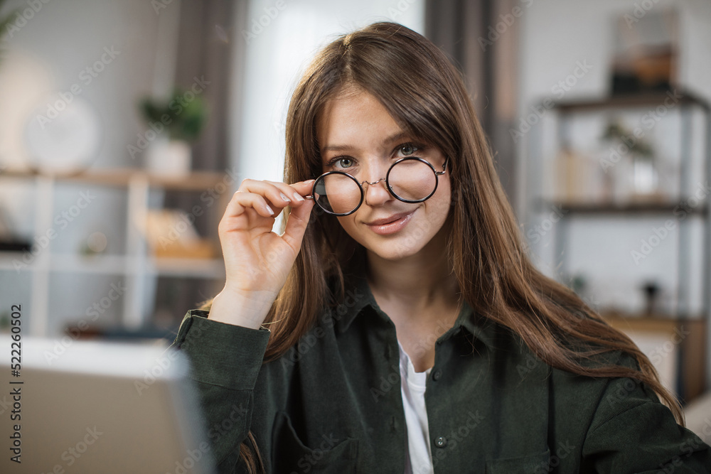 Pretty young caucasian woman with long dark hair sitting at table with modern laptop and touching glasses with hands. Concept of freelance and technology. Pretty young caucasian woman with long dark hair sitting at table with modern laptop and touching glasses with hands. Concept of freelance and technology.
