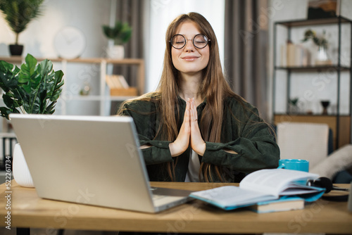 Relaxed caucasian woman in casual wear sitting on workplace with modern laptop and meditating with closed eyes. Office worker relief street at work with accomplished fingers.