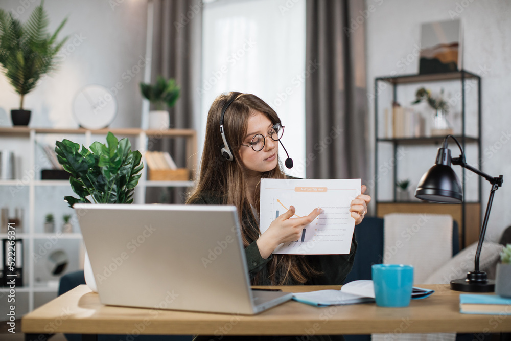 Caucasian woman presenting financial report to colleagues through video call on laptop. Female freelancer sitting at workplace and talking in headset. Caucasian woman presenting financial report to colleagues through video call on laptop. Female freelancer sitting at workplace and talking in headset.