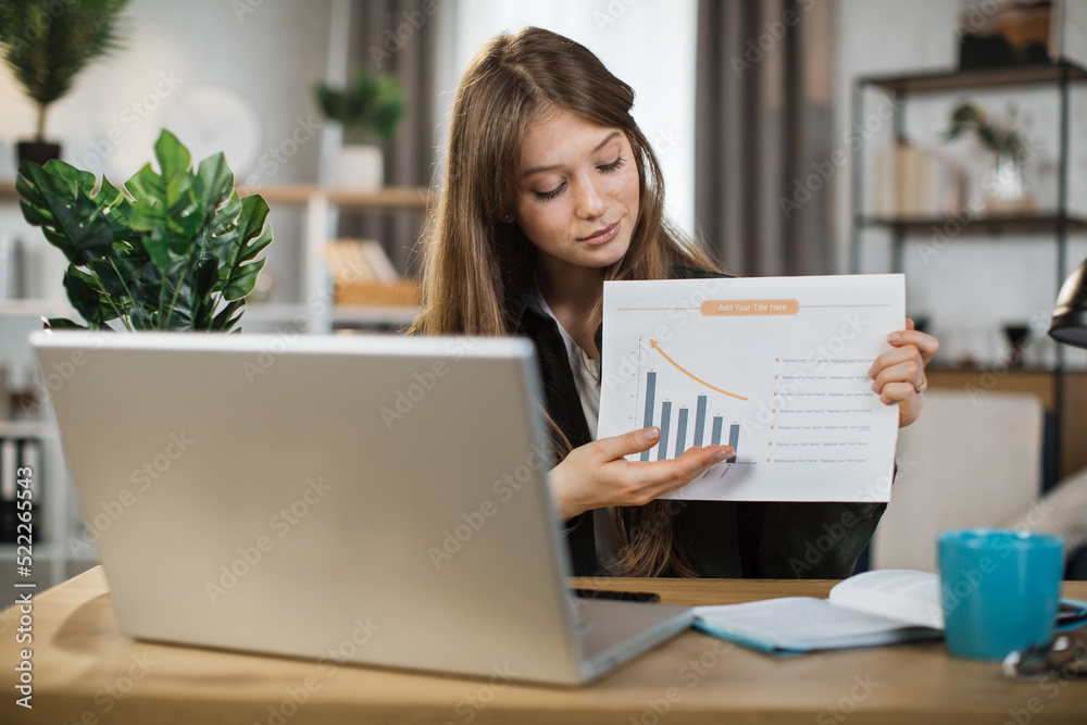 Smiling female manager leading working meeting through video call on laptop. Young woman freelance showing graphs and chart while sitting at table. Smiling female manager leading working meeting through video call on laptop. Young woman freelance showing graphs and chart while sitting at table.