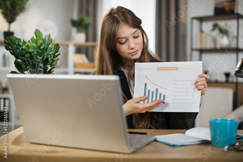 Smiling female manager leading working meeting through video call on laptop. Young woman freelance showing graphs and chart while sitting at table.