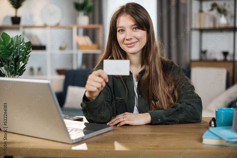 Young pretty woman with casual outfit sitting at desk with wireless laptop and showing empty white card. Space for text on business card. Young pretty woman with casual outfit sitting at desk with wireless laptop and showing empty white card. Space for text on business card.