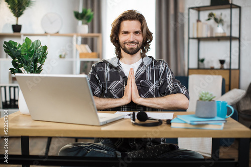 Young bearded man freelancer sitting at table with opened eyes and relieving stress by meditation at workplace. Concept of relaxation and harmony, no stress free relief at work.