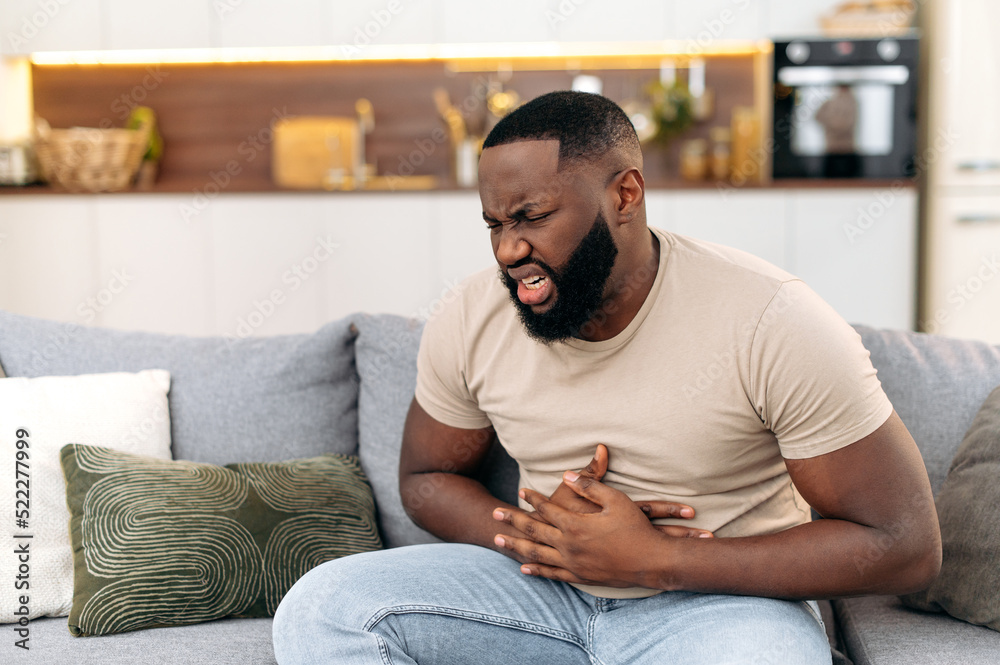Unhappy guy of african american ethnicity, sits on a sofa in the living room, holds his hands on his stomach, grimaces from pain in his stomach, suffers from poisoning, spasm, stomach problems