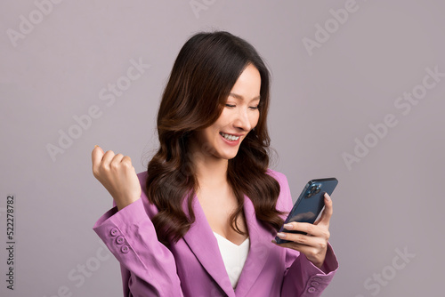 Asian woman with long hair, wearing a pink suit, holding a smartphone, happy gestures on gray background.