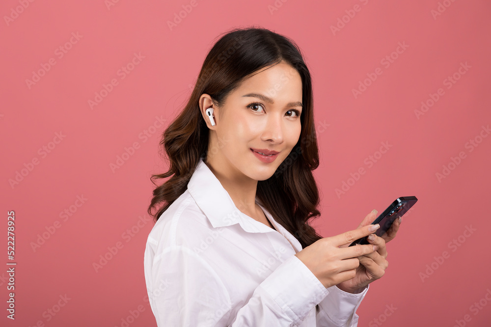 long hair Asian woman wearing a white shirt Holding phone and wearing wireless earphones on a pink background. long hair Asian woman wearing a white shirt Holding phone and wearing wireless earphones on a pink background.