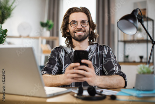 Confident young man writing message during mobile conversation at office. Caucasian male worker with long dark hair sitting at table with opened laptop.