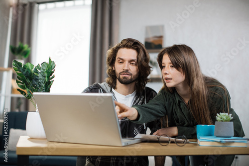 Young man and woman in casual outfit looking at computer screen during working meeting. Young business partners cooperating at bright office.