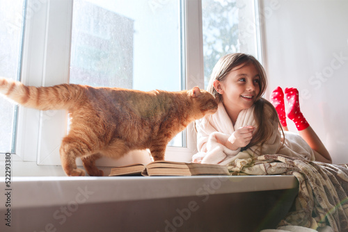 Little girl with red cat lying on window sill , reading a book Autumn weekend with cat at home.