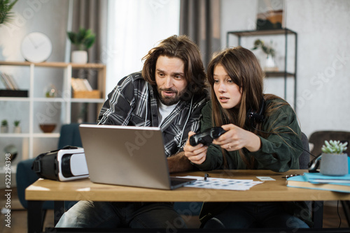 Young happy couple playing video games at home, sitting at the table with laptop and using joysticks. Happy man in headset shows new game for his lovely smiling girlfriend at home.